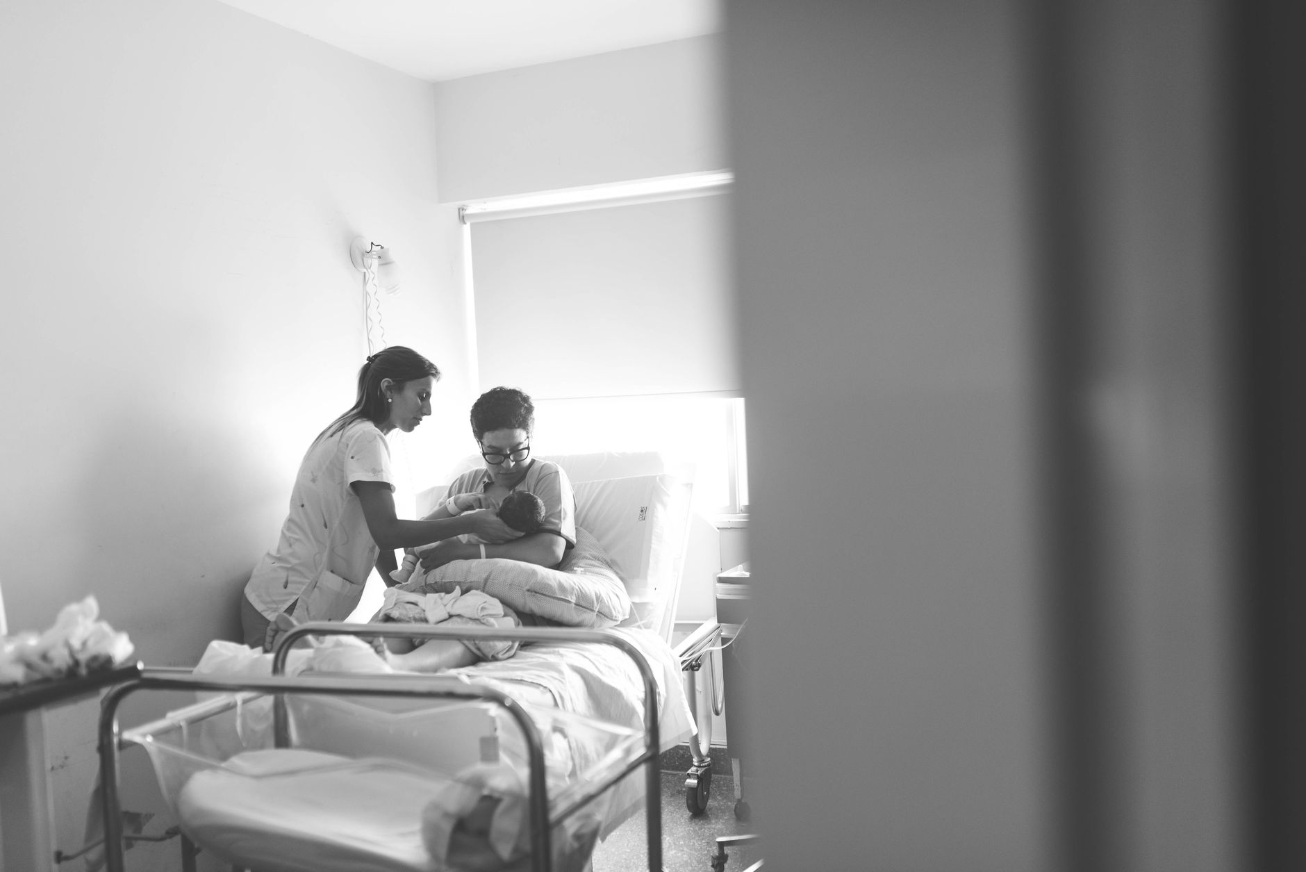 A calm maternity ward room with a supportive nurse holding a newborn, soft natural light from a window, pastel curtains, and a bedside table with a bottle of water and a pill bottle