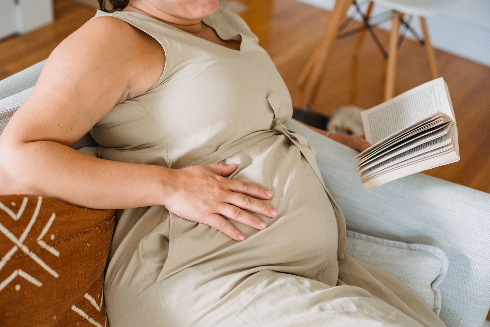 A pregnant woman in a cozy living room, seated on a soft sofa, sipping herbal tea while reading a pregnancy guide