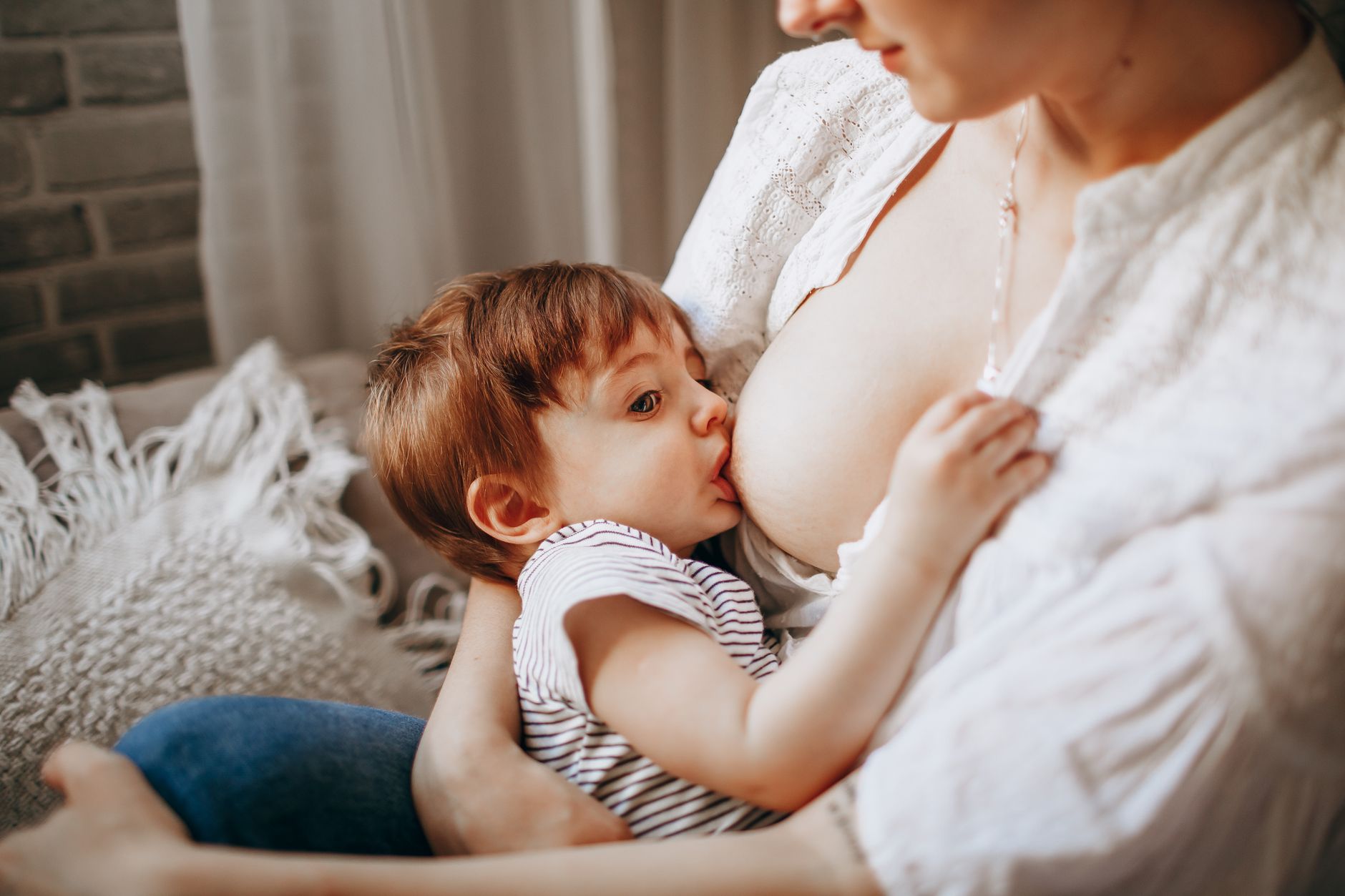 A mom sitting comfortably on a couch, using a nursing pillow to support her back while breastfeeding her baby