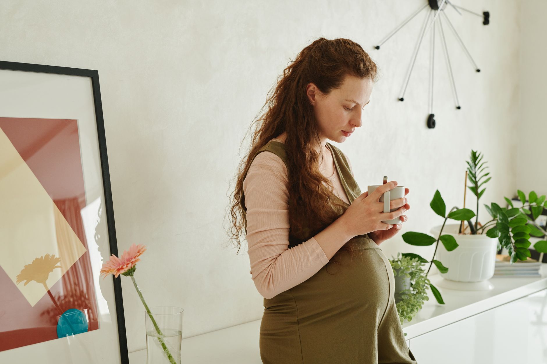 A calm living room scene with a pregnant woman holding a warm cup of tea, soft blanket draped over a sofa, gentle sunlight filtering through curtains, pastel tones, photorealistic, high detail