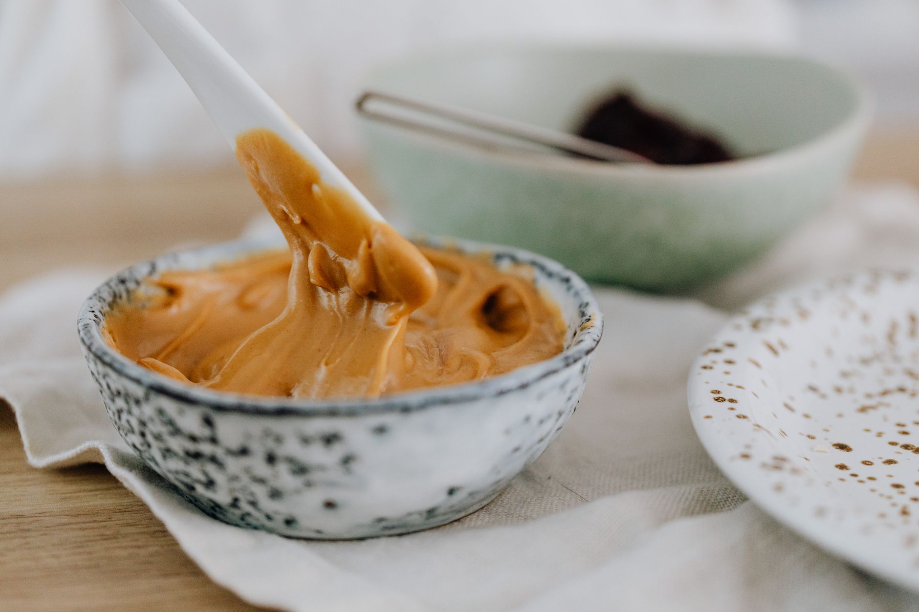 A close‑up of a spoonful of thinned peanut butter on a pastel baby plate, with a soft‑focus kitchen background, warm morning light, inviting and safe