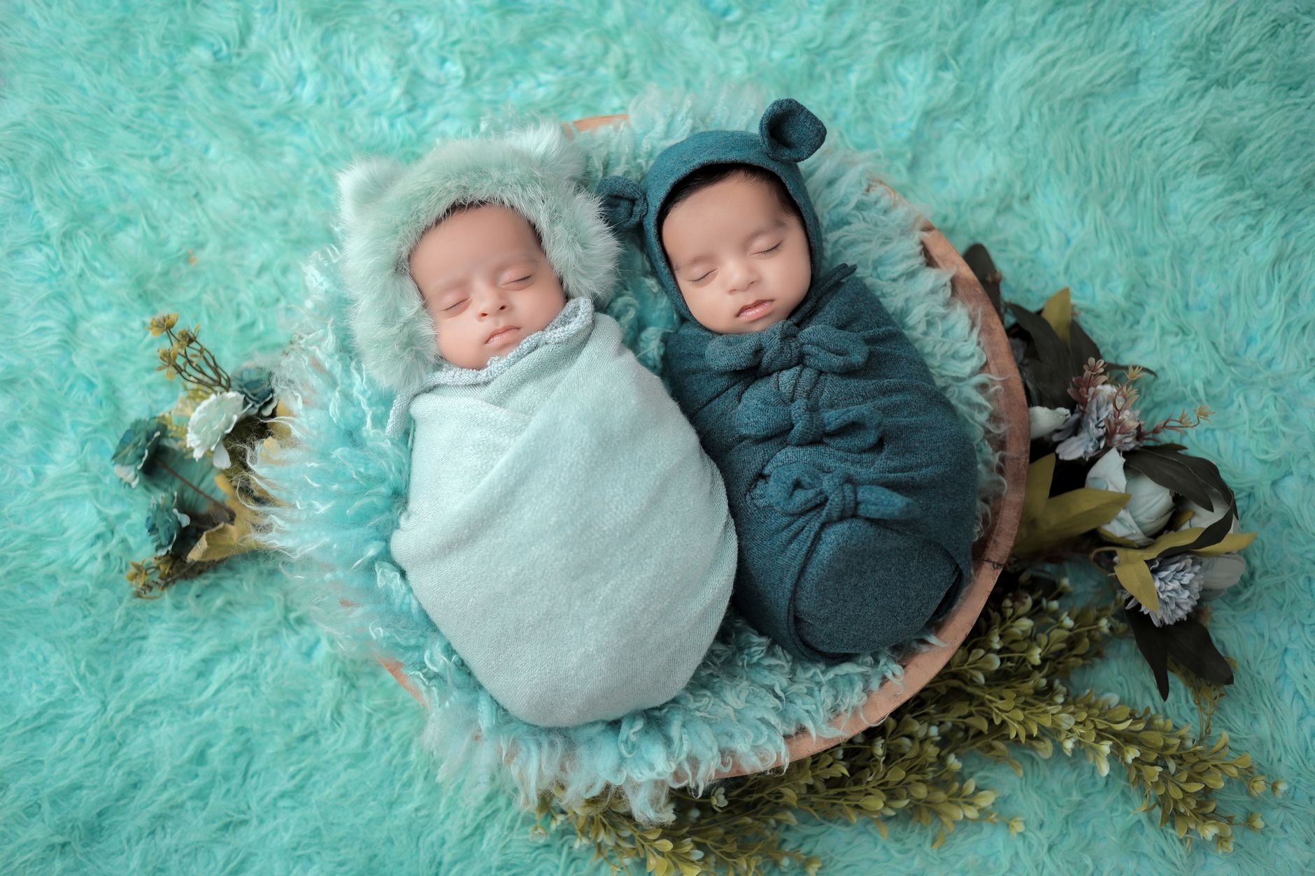 Twin newborns swaddled side by side on a soft blanket, with a sleepy mother reaching out in the background