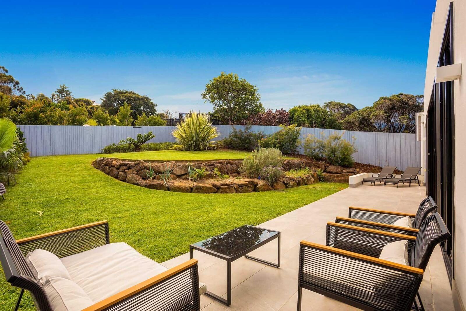 Level rear backyard with a green lawn, curved rock retaining wall, and an outdoor patio with seating.
