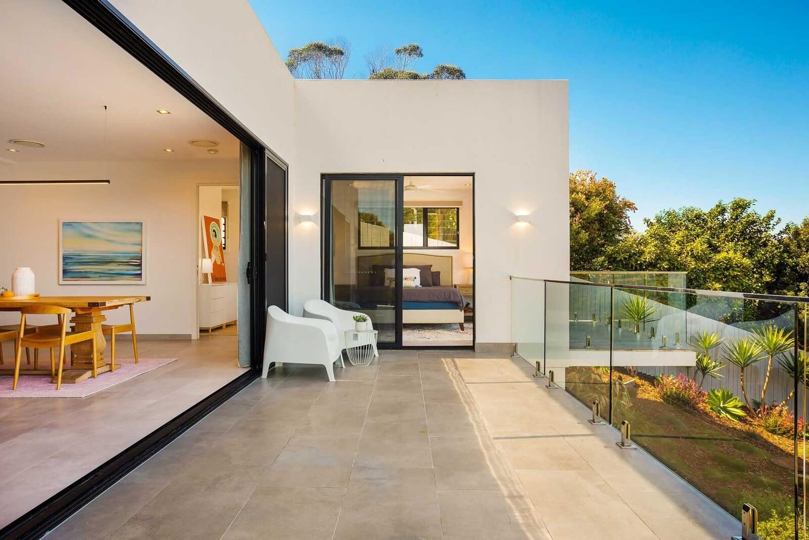 Tiled outdoor balcony with a glass balustrade and white outdoor chairs, connecting to an open bedroom and living space.