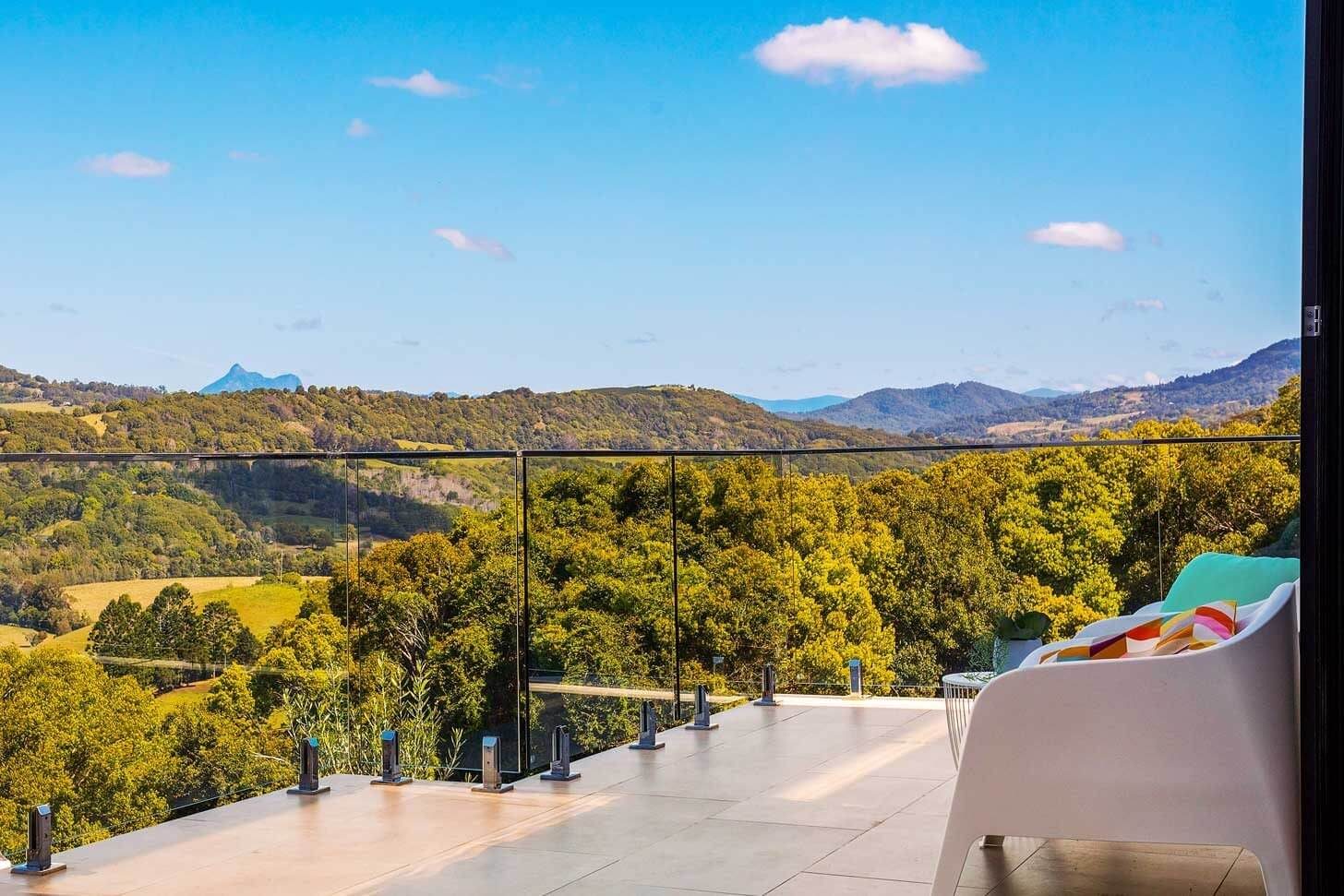 Perspective from an elevated tiled balcony with a frameless glass balustrade overlooking a vast, lush green valley and rolling hills under a bright blue sky.