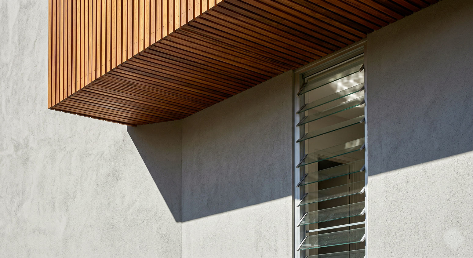 Architectural detail of a deep timber-lined eave casting a sharp diagonal shadow across a rendered wall, with a tall louvre window for cross-ventilation.