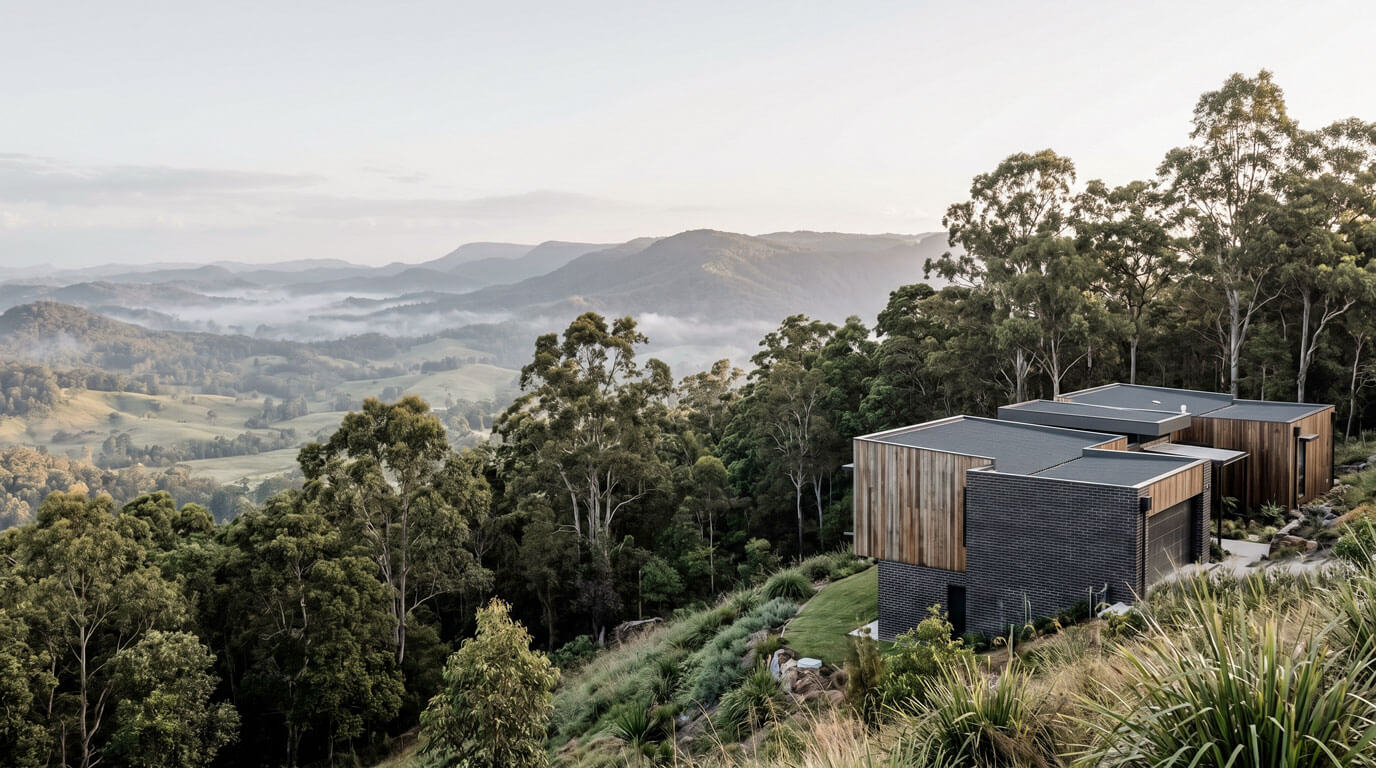 A contemporary home of dark brick and timber pavilions sitting on a hinterland ridge, looking out over a misty valley framed by eucalyptus trees at dawn.