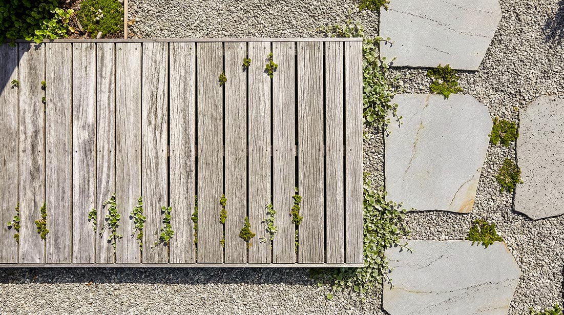 A top-down photograph of a path where a section of weathered silver-grey timber boardwalk planks meets large, irregular flagstone pavers set on a bed of fine gravel. Small green groundcover plants grow through the gaps and edges.