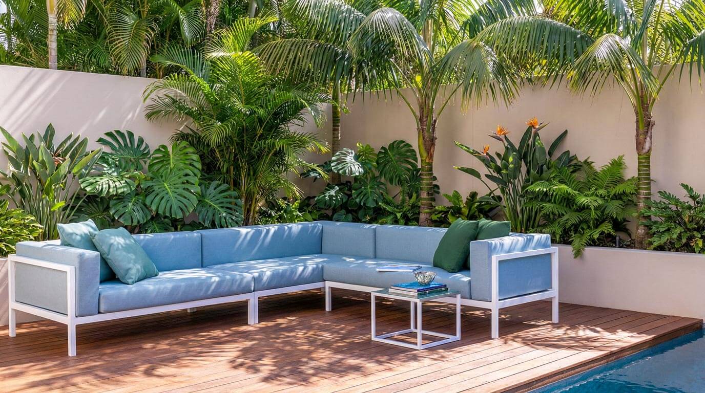 A contemporary blue outdoor sectional sofa resting on a timber pool deck, framed by a low rendered planter wall and lush, architectural subtropical plants including Monstera and palms.