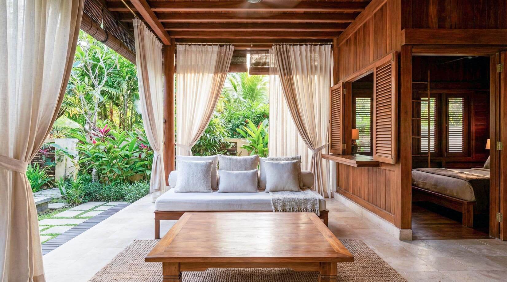 An intimate outdoor lounge area under a timber pergola with retractable canvas canopy shades, set against a vertical timber batten privacy screen and layered, broad-leaf tropical foliage.