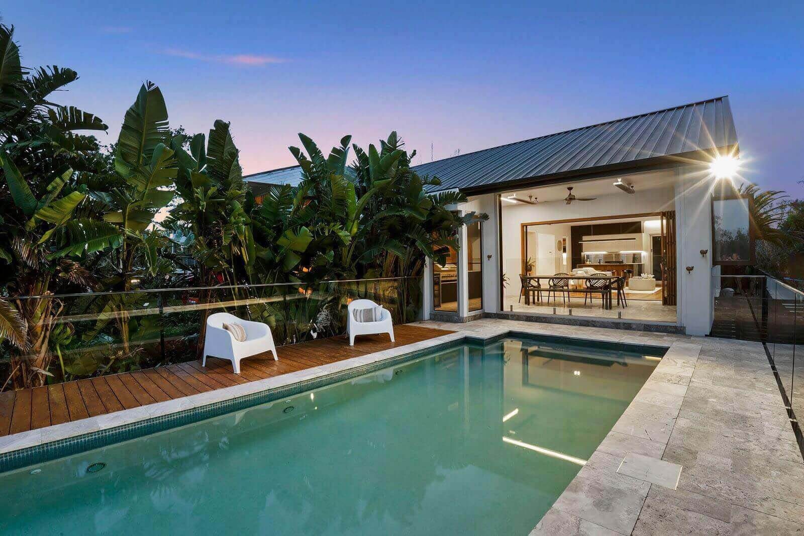 Twilight shot of a backyard pool and deck with lush tropical landscaping and the house illuminated in the background.