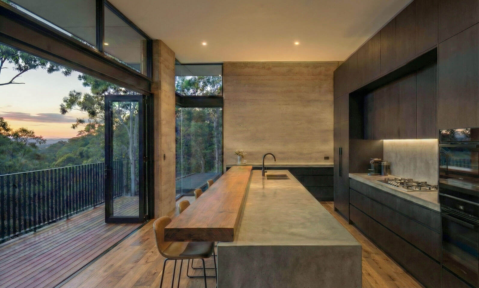 Modern kitchen interior featuring a long off-form concrete island bench with a timber extension. Dark cabinetry lines the wall. Large glass doors open onto a timber deck with views of the sunset and gum trees.