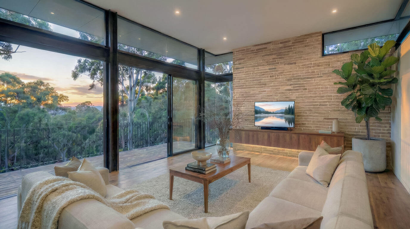 Living room featuring a textured face-brick internal wall and timber flooring. Floor-to-ceiling glass sliding doors frame a view of the native tree canopy. Neutral furniture sits on a textured rug.