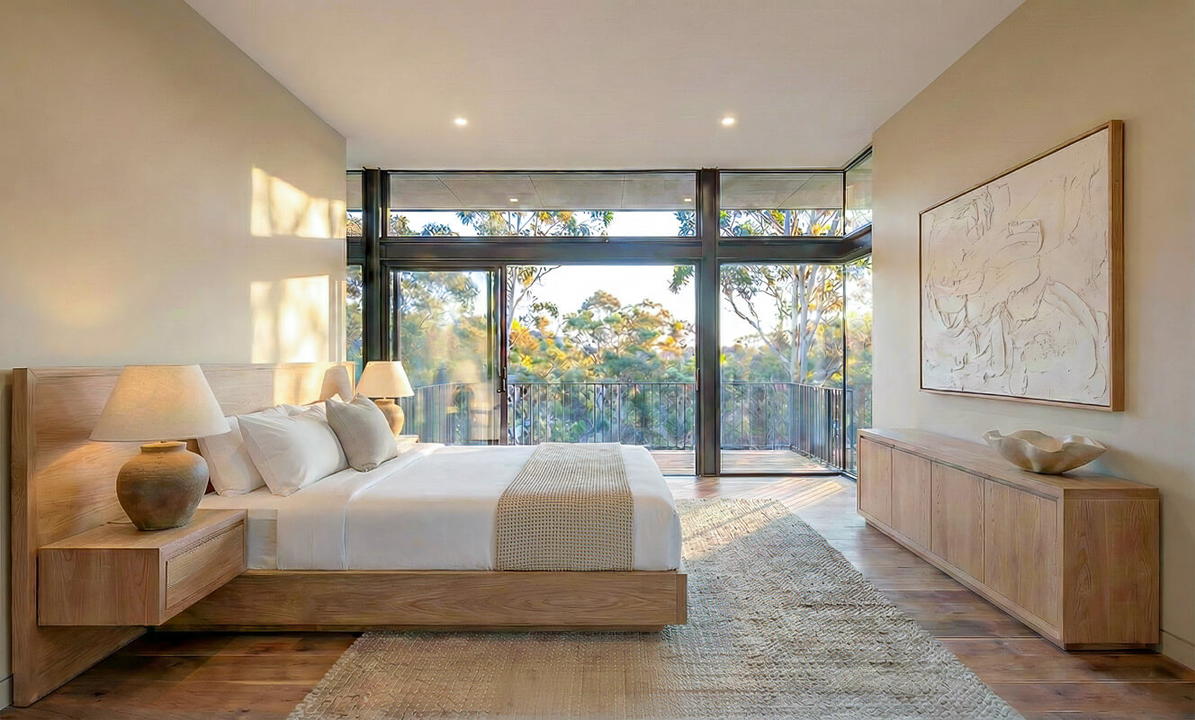 Master bedroom with a timber bed frame and side tables. A corner window frames the native bushland. Soft morning light casts shadows on the beige walls and textured artwork.