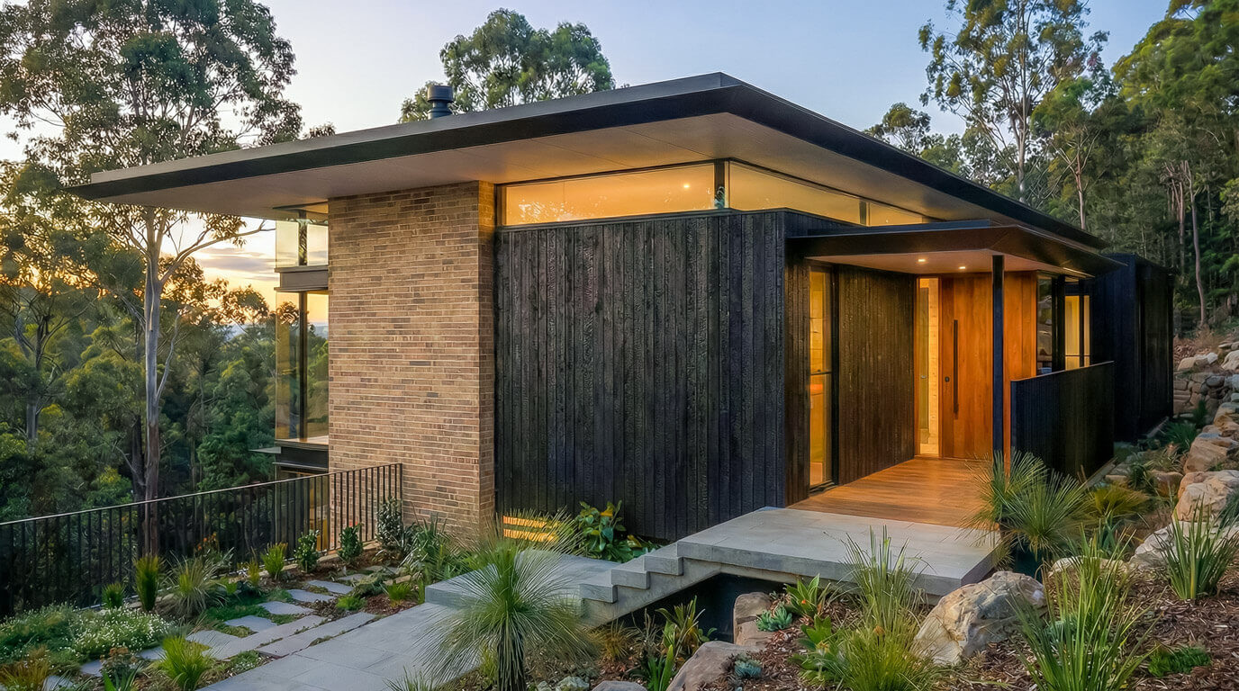 Front facade of Ironbark Ridge at twilight. The home features a flat, cantilevered roof, Shou Sugi Ban charred timber cladding, and a warm timber entry door. Native stone landscaping surrounds the concrete steps.