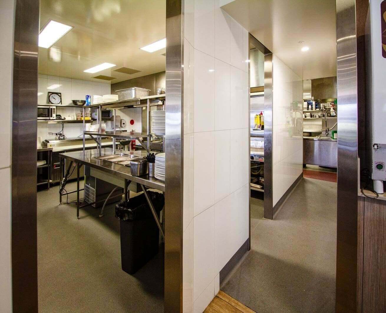 A view through a stainless steel portal into a professional kitchen featuring metal prep benches and industrial shelving.