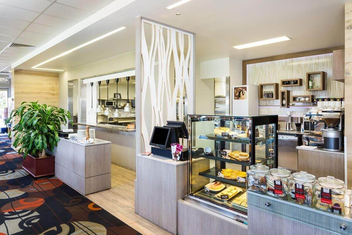A cafe service counter with a glass pastry display case, light timber-look panelling, and a decorative white laser-cut screen.