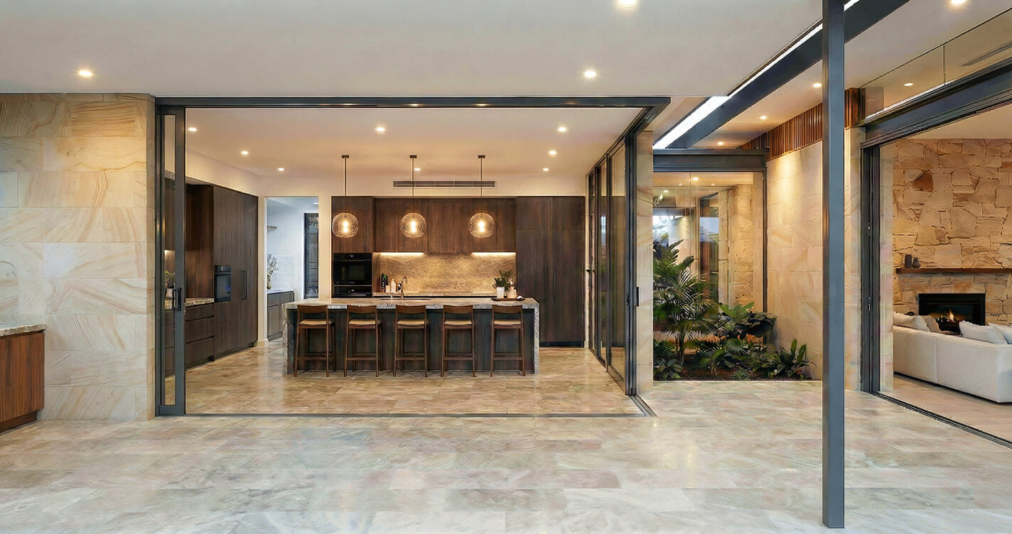 Modern kitchen with dark timber cabinetry and a stone island bench. Large glass sliding doors are open, connecting the kitchen directly to a covered alfresco dining area with sandstone walls.