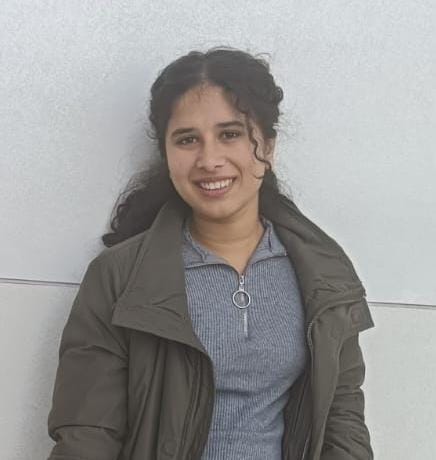 A smiling young woman with dark, wavy hair styled half-up, wearing an olive-green jacket over a grey ribbed zip-up top