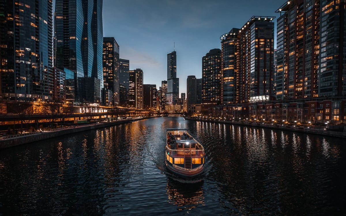 Decorated and illuminated boats parade along the Chicago lakefront at night with the city skyline glowing behind them.