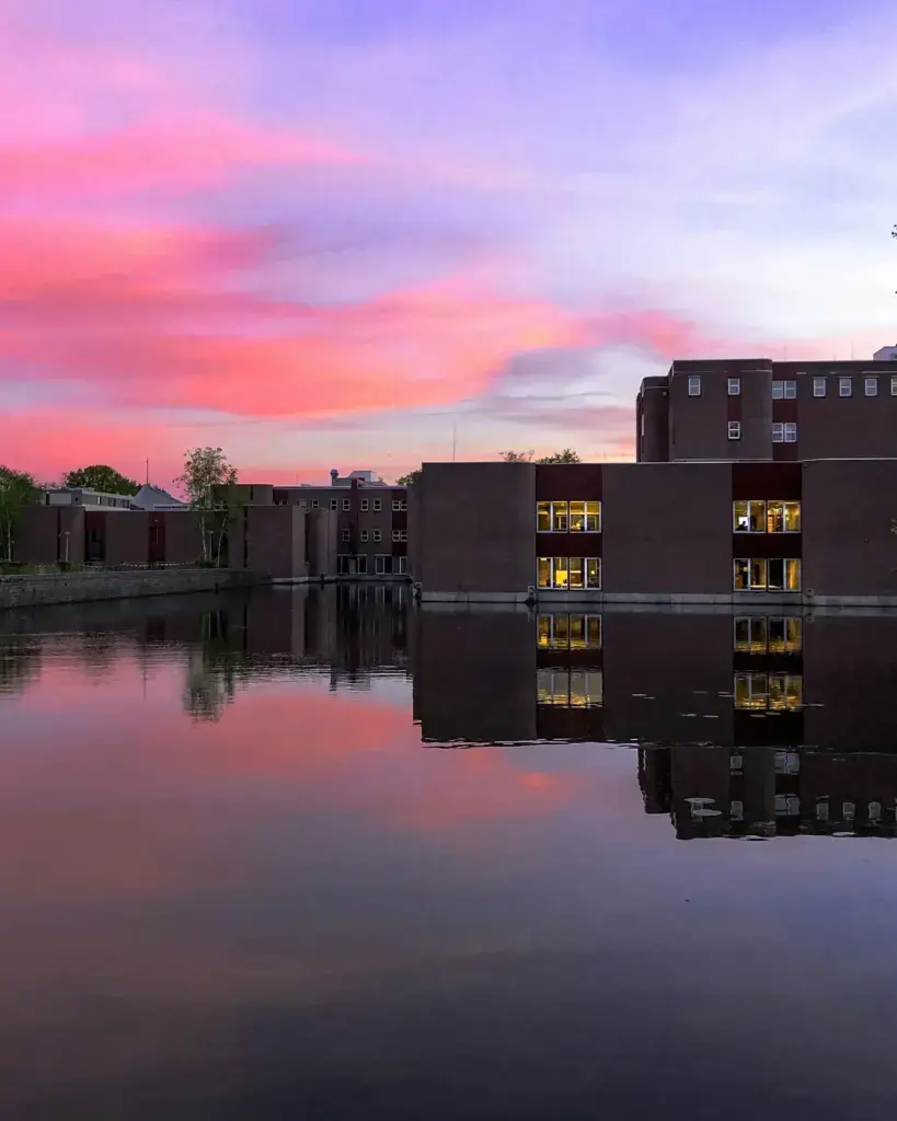 Modern dark-brick building with lit windows mirrored in still water at sunset in Enschede, under a pink and purple sky