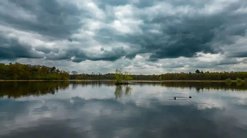 Still lake water at the Galderse Meren near Breda reflecting a heavy grey cloudy sky, with a wooded shoreline in the distance