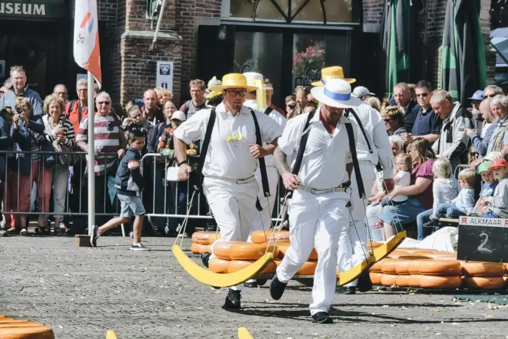 Two cheese carriers in white uniforms and straw hats running with a yellow barrow of cheeses across the Waagplein in Alkmaar, watched by a crowd behind barriers