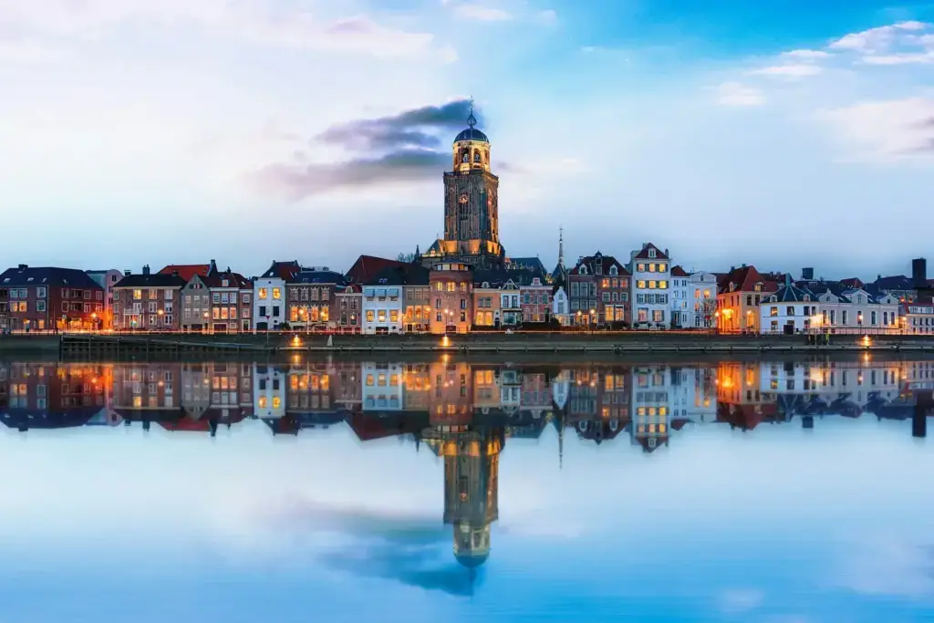 Deventer skyline reflected in the IJssel at dusk, with the lit Lebuinus church tower rising above the row of quayside houses