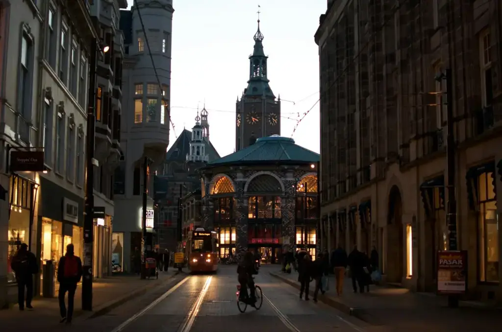 Shopping street in The Hague at dusk with tram tracks, a cyclist and the tower of the Grote Kerk rising behind the rooftops