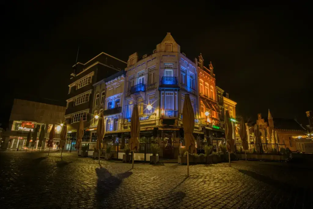 Market square in Den Bosch at night, with a corner building lit in blue and orange and closed café parasols on the wet cobbles
