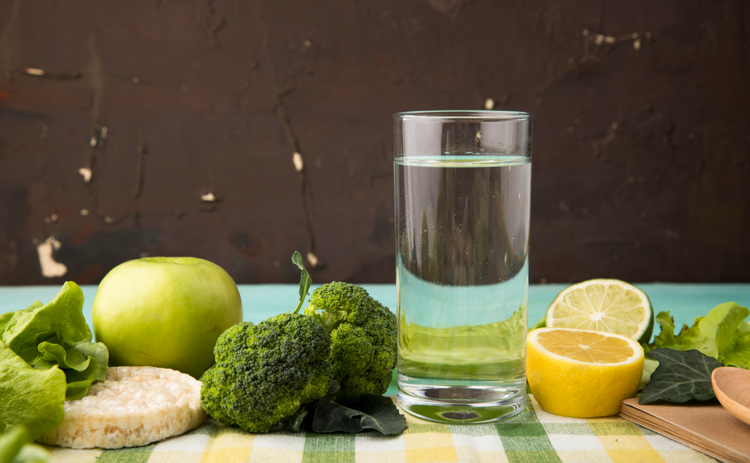A glass of water with fresh fruits and vegetables around it.