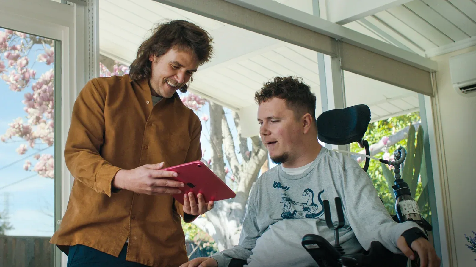 Two people looking at a tablet together, one seated in a powered wheelchair.