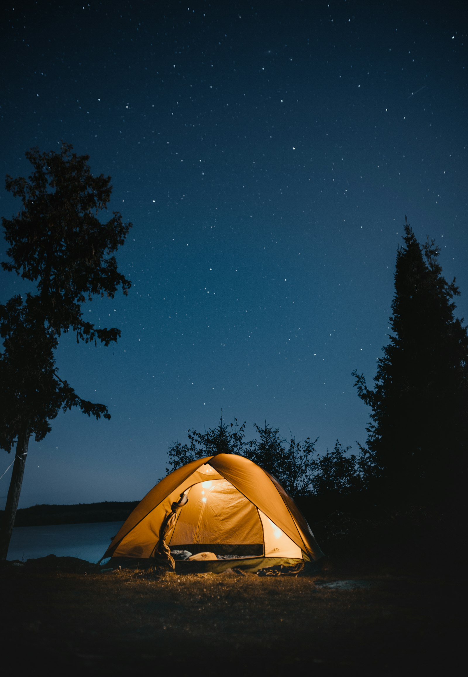A lit yellow camping tent under a starry night sky beside water and trees