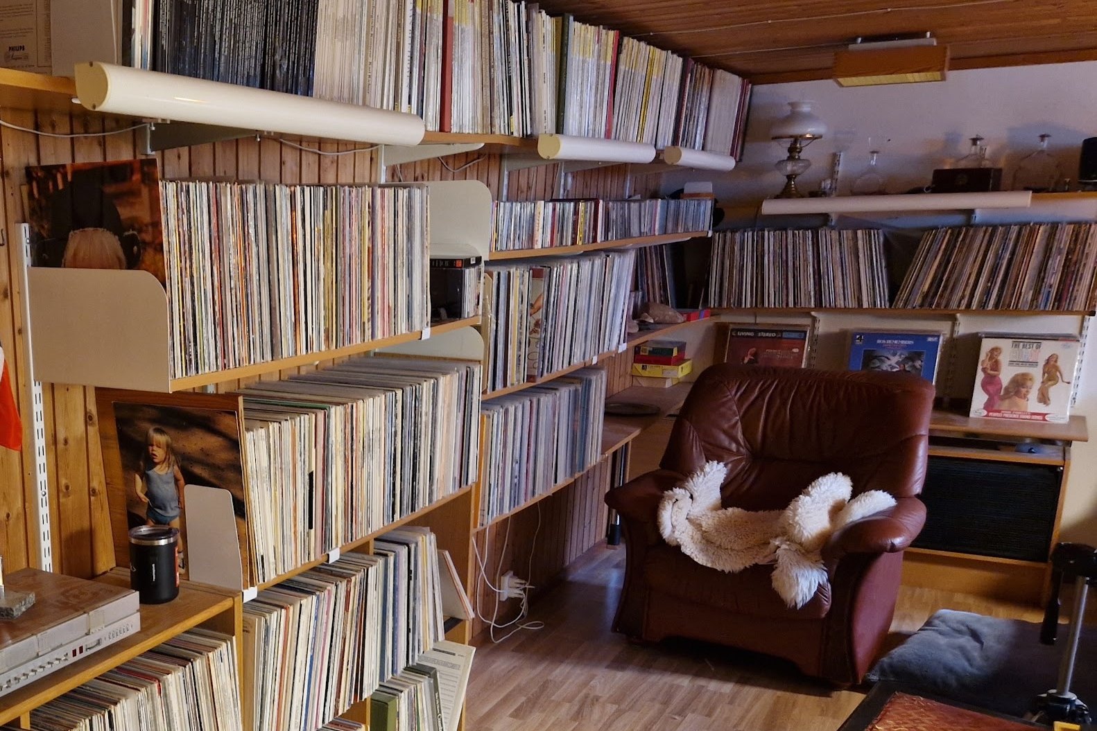 A home record room with shelves of LP records along the walls, a brown leather armchair with a sheepskin throw and more shelves of records behind it
