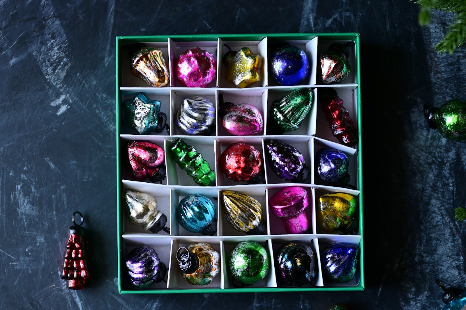 A green box of vintage glass ornaments in white compartments, with a few baubles on a dark table