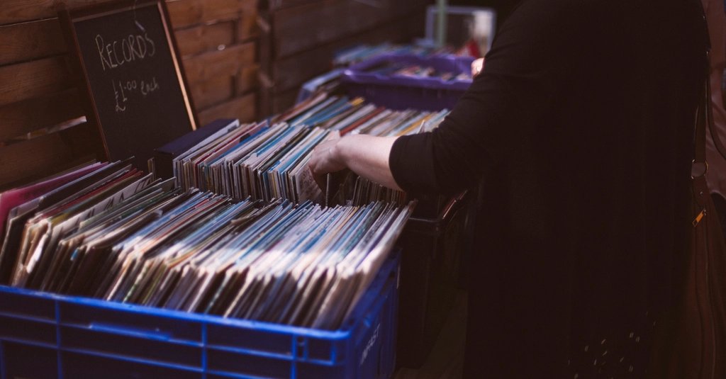 A hand flips through crates of second-hand vinyl records at a market stall, beside a chalkboard reading Records, one pound each