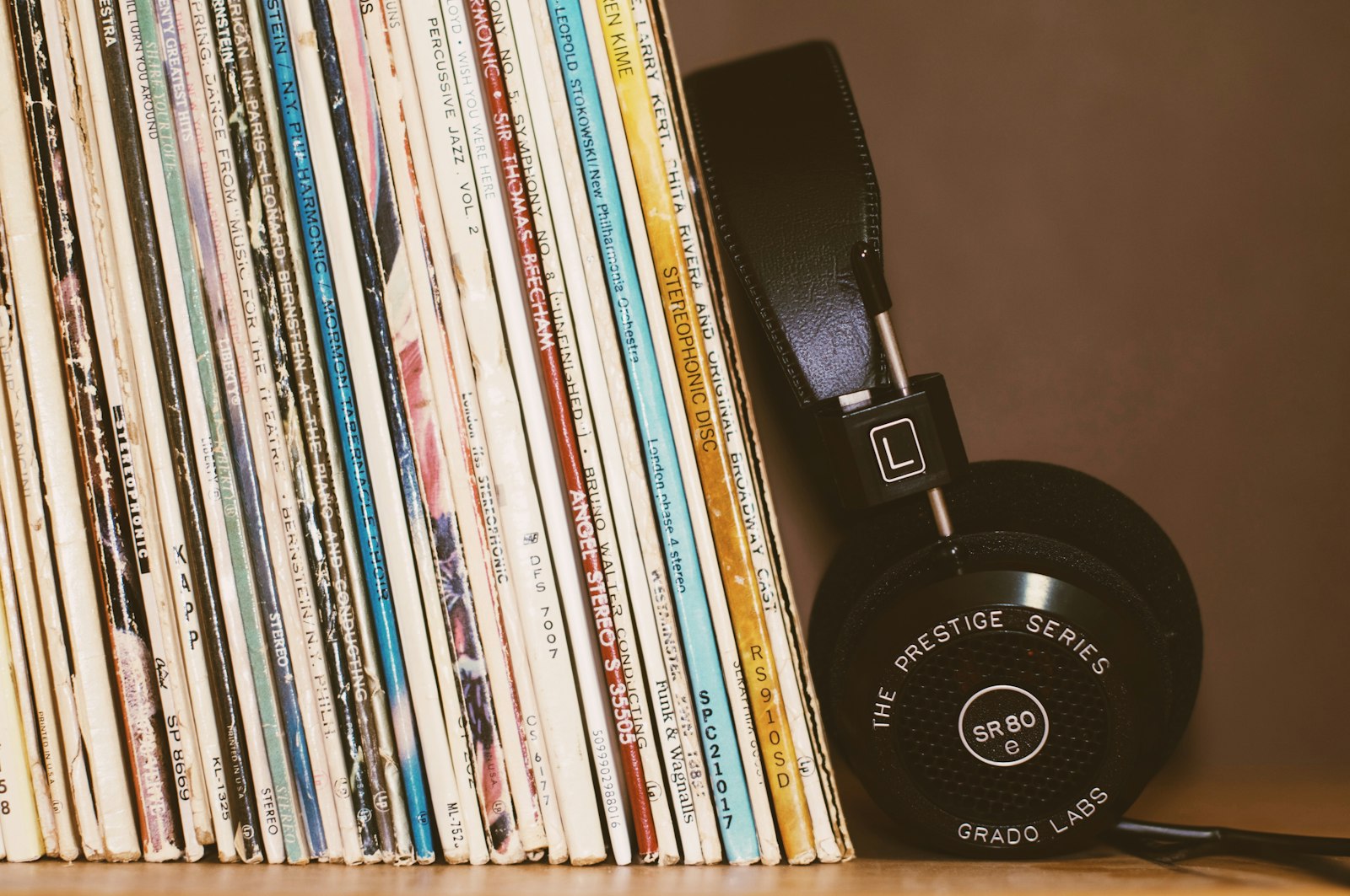 A row of worn vinyl record sleeves on a wooden shelf with black headphones resting against them