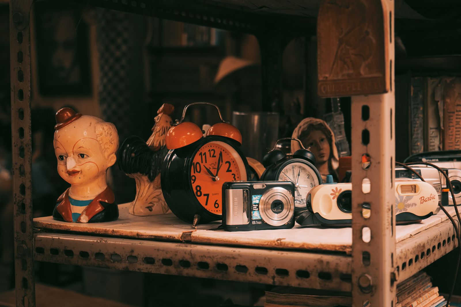 A shelf crowded with mismatched objects in warm light: two alarm clocks, a ceramic rooster, an old doll head, a compact camera and a toy camera, with books stacked behind them