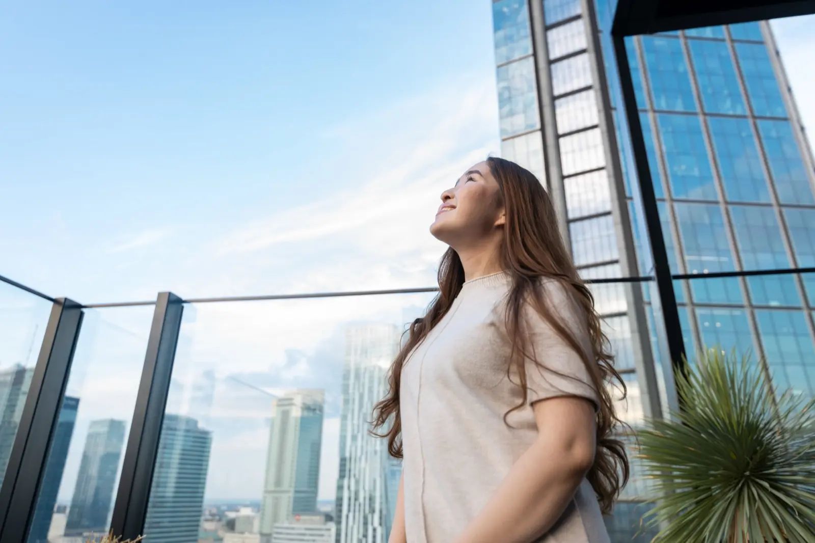 A woman on a city rooftop terrace gazing up at the sky between tall glass buildings