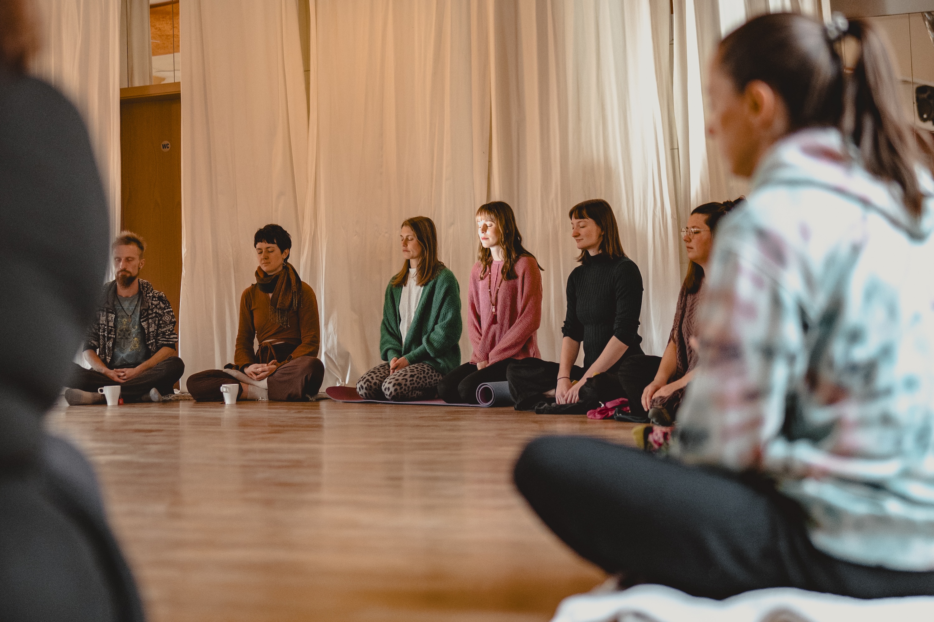 Mehrere Menschen sitzen zusammen im Opening Circle. Man sieht einen Teil des Kreises. Die Sonne scheint dabei sanft in die Gesichter einzelner Teilnehmer*innen. 