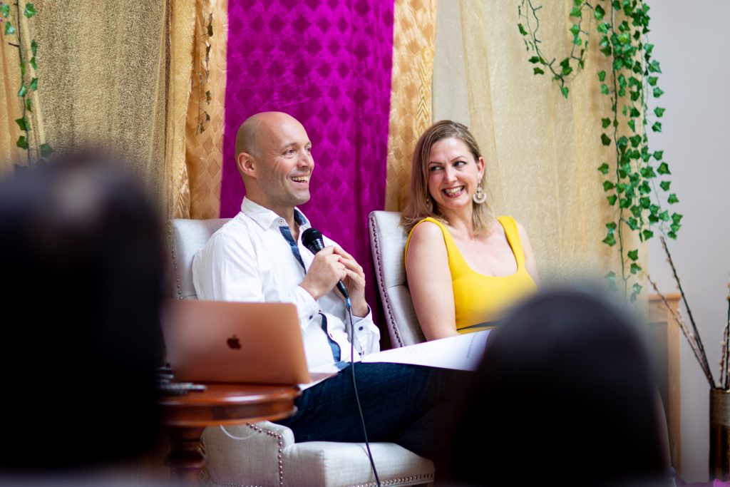 Two teachers smiling on stage during a talk, one holding a microphone