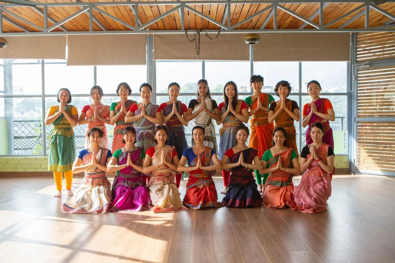 A group of women in colorful attire posing with their hands in a prayer position inside a Yoga.