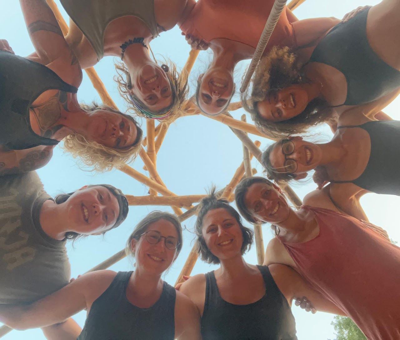 A group of women smiling and posing in a circular arrangement under a wooden structure.