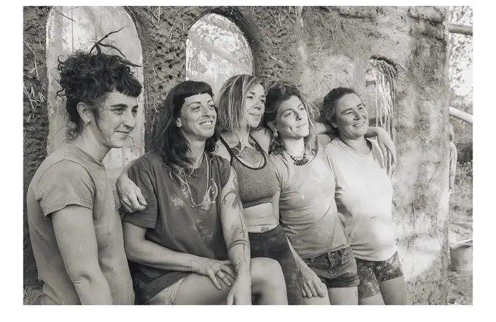 A group of five women posing together in front of a mud wall.