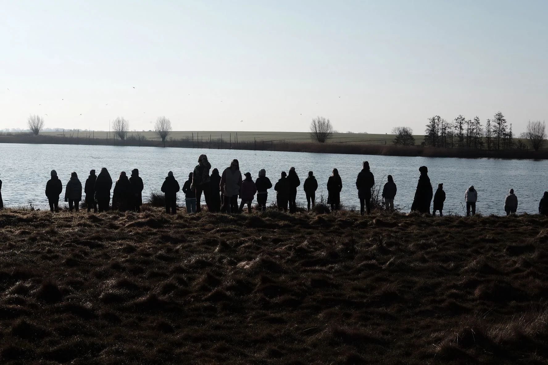 Das Bild zeigt eine Gruppe von Menschen, die silhouettenhaft am Ufer eines Gewässers stehen.