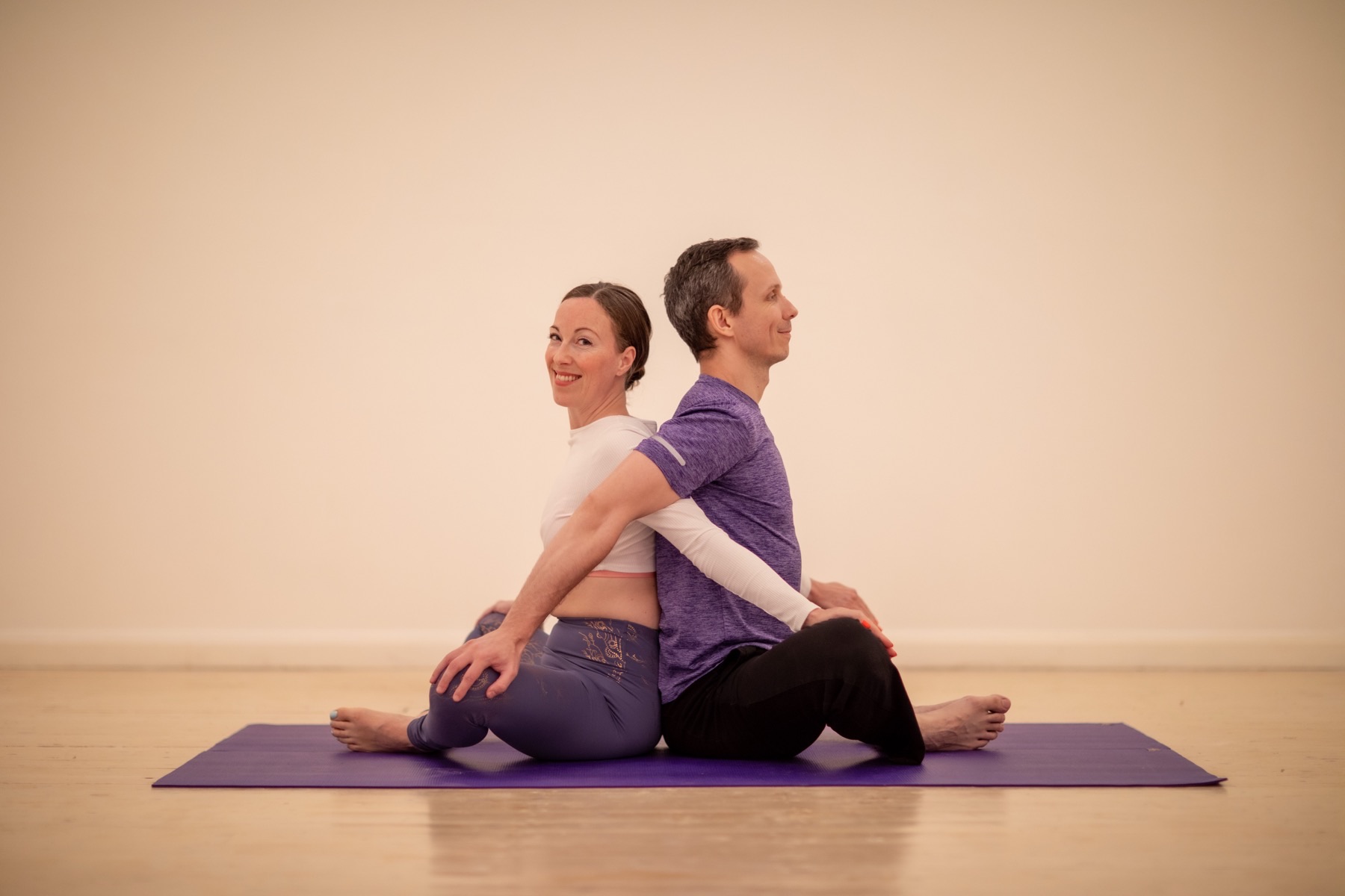 A woman and a man sitting back to back on a yoga mat, smiling