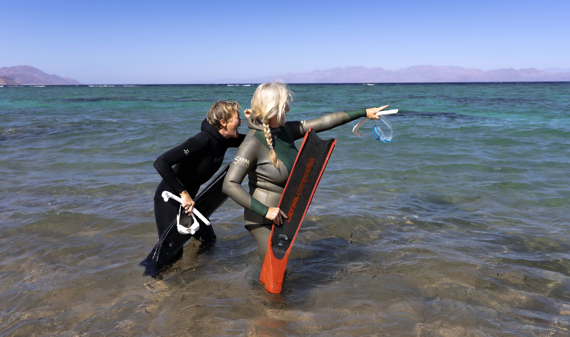 Zwei Personen stehen im Wasser und bereiten sich auf das Schnorcheln vor, eine zeigt auf etwas im Wasser.