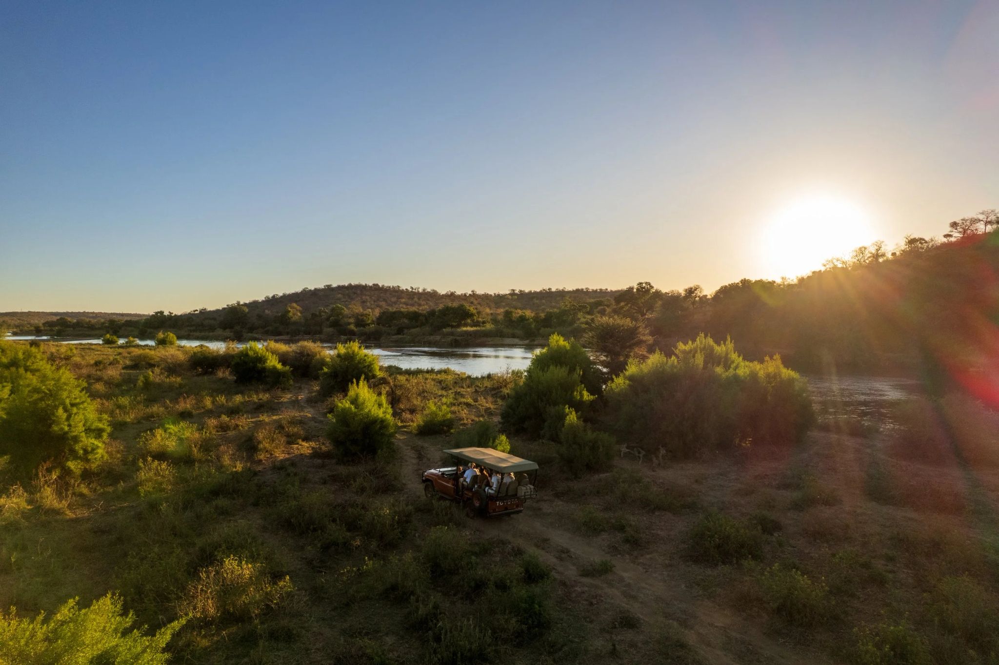Safari vehicle parked along the riverbank at sunset in Greater Kruger National Park, surrounded by golden light and wild bushland.