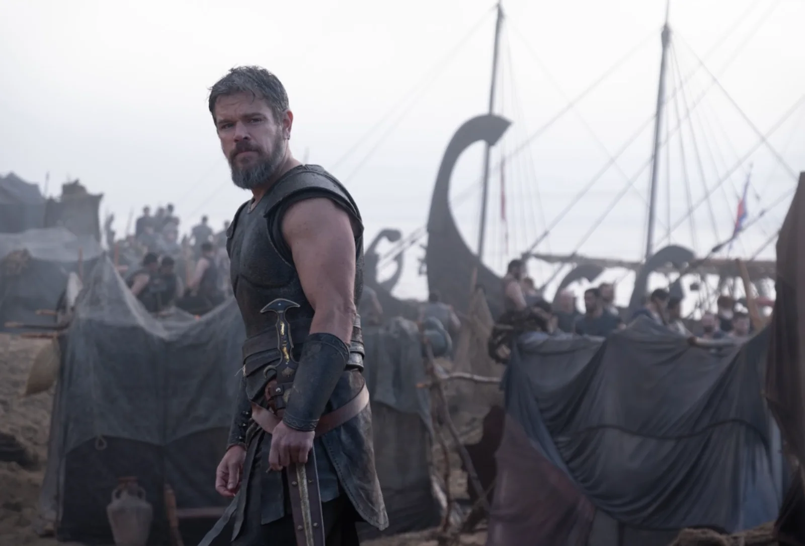 Matt Damon como Aquiles en Troya con armadura y espada, barcos de guerra al fondo