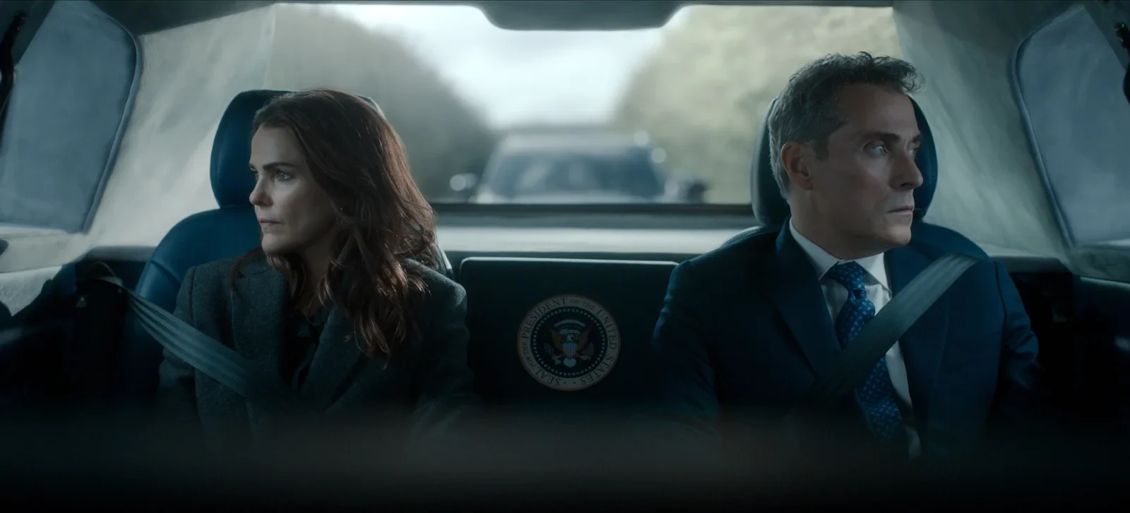 Keri Russell y Rufus Sewell sentados en el asiento trasero de un auto oficial, en La diplomática